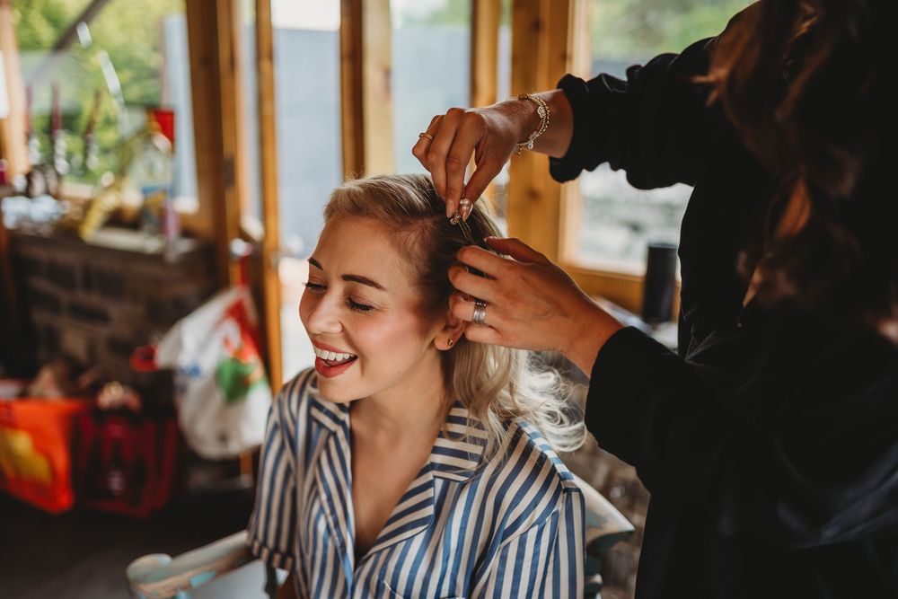 Smiling bride in a striped robe has her hair styled by a hairdresser in a sunlit room while getting ready for her wedding.