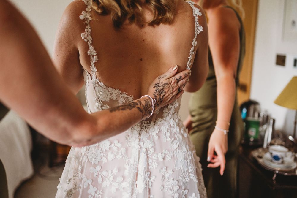 Bride in a lace wedding gown with an open back as a helper adjusts the dress and fastens the buttons before the ceremony.