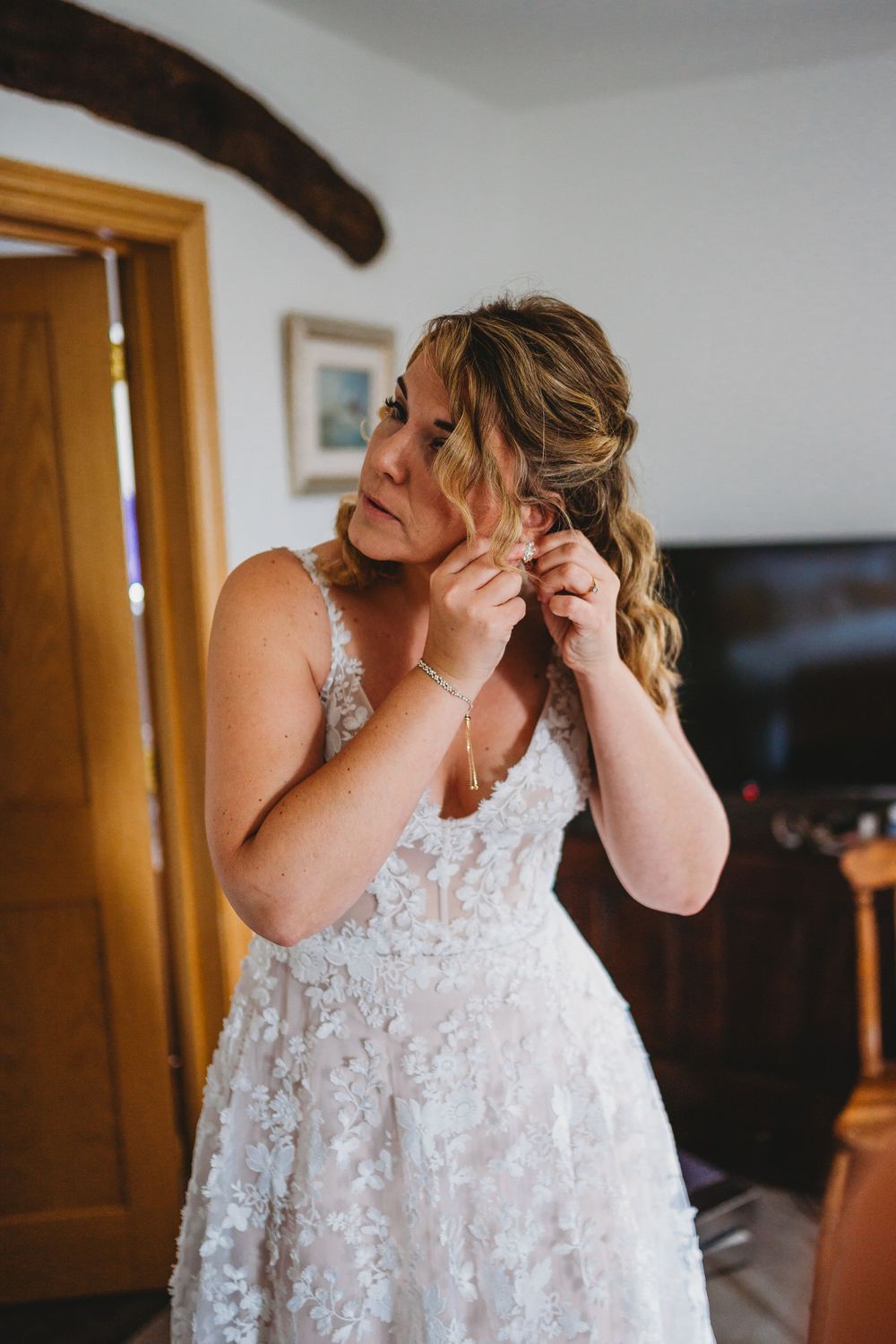 Bride in a lace floral wedding gown with wavy half-up hair adjusts her earring in a bright room before the ceremony.
