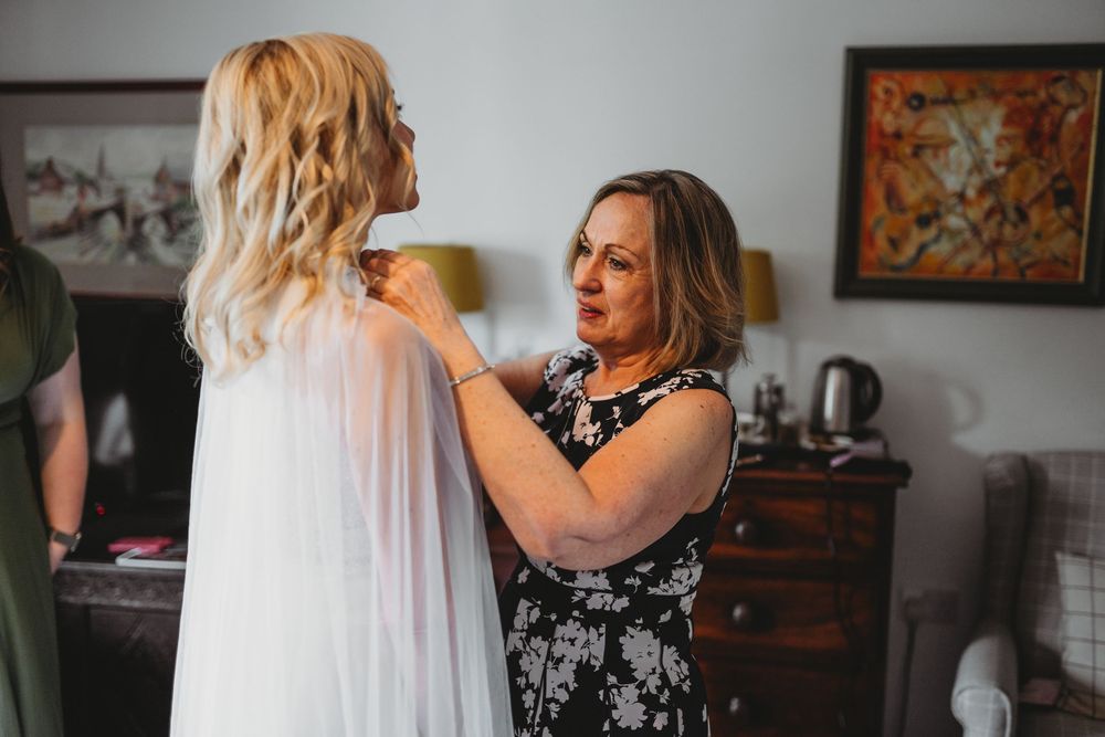 Older woman in a floral dress helps a blonde bride in a sheer veil adjust her wedding attire in a cozy room before the ceremony.