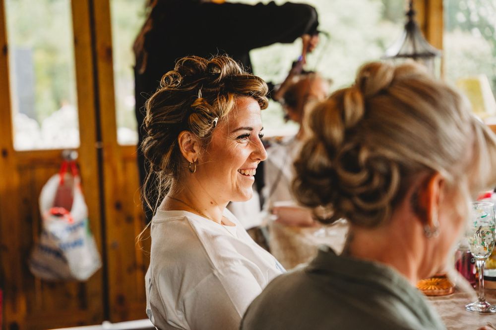 Smiling woman with an elegant braided updo getting ready in a white robe while a stylist works behind her in a rustic wooden room.