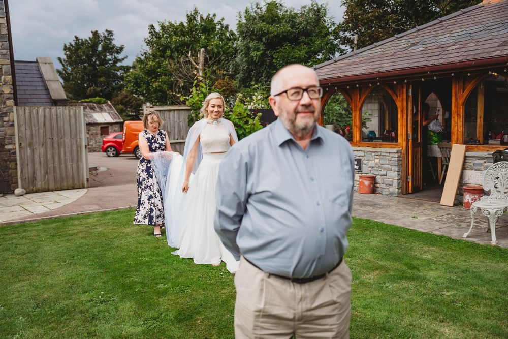 A smiling bald man with glasses stands in the foreground while a bride in a white gown is helped by a bridesmaid on a lawn behind him.