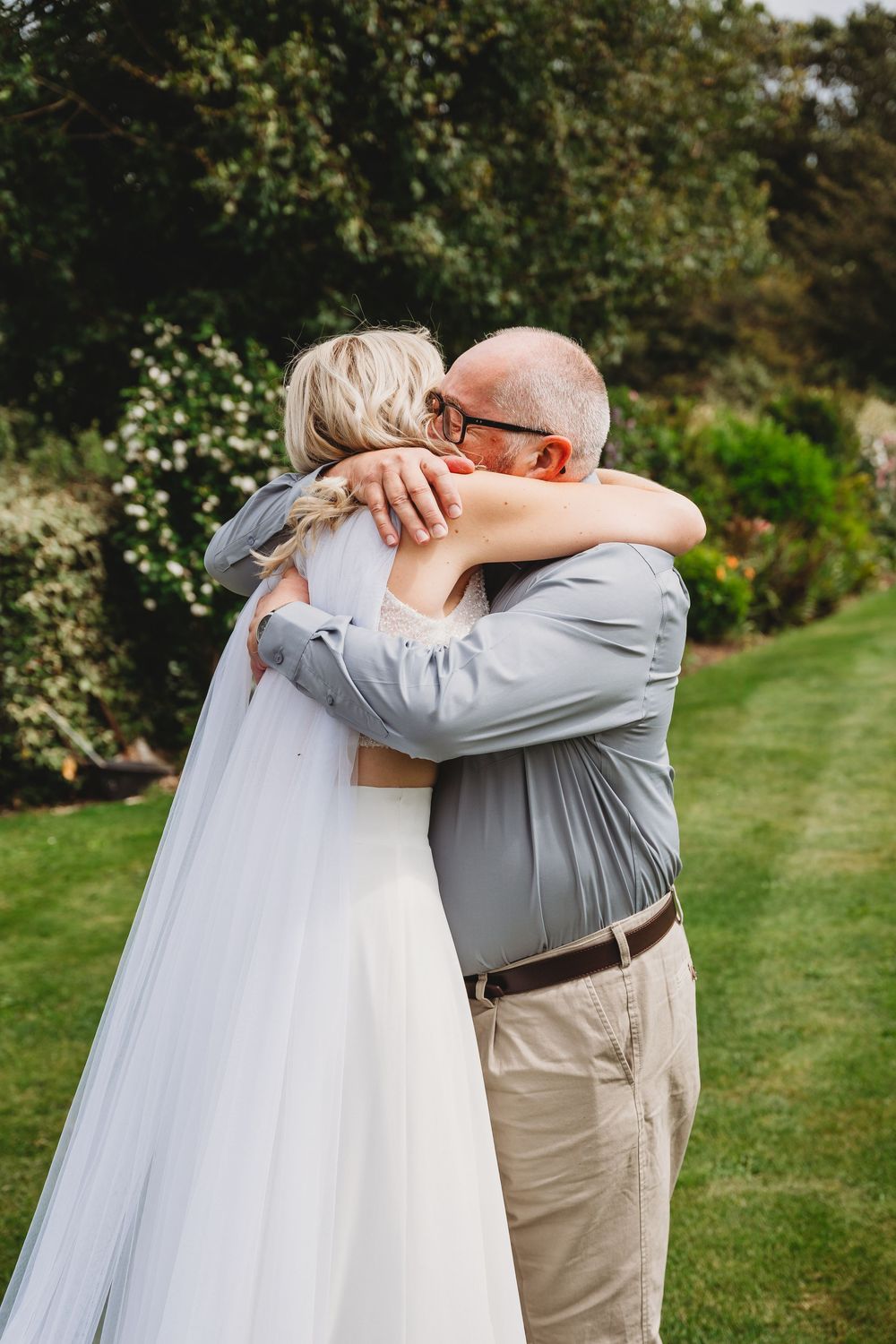 A bride in a white wedding dress and long veil shares a heartfelt embrace with an older man in glasses in a lush green garden.