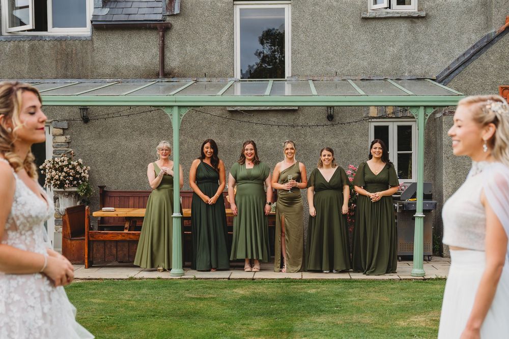 Six bridesmaids in olive green dresses stand under a glass-roofed pergola on a lawn as two women in white dresses face them.