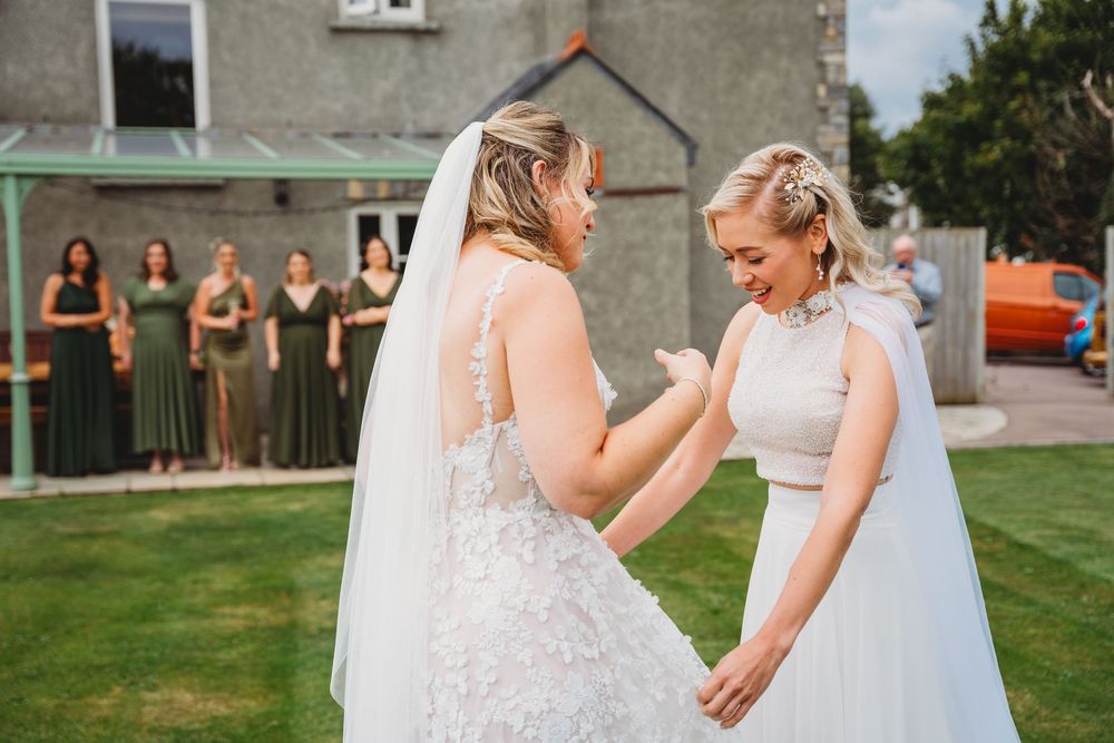 Two brides in white wedding dresses with veils hold hands on a lawn while bridesmaids in green dresses watch from a patio.