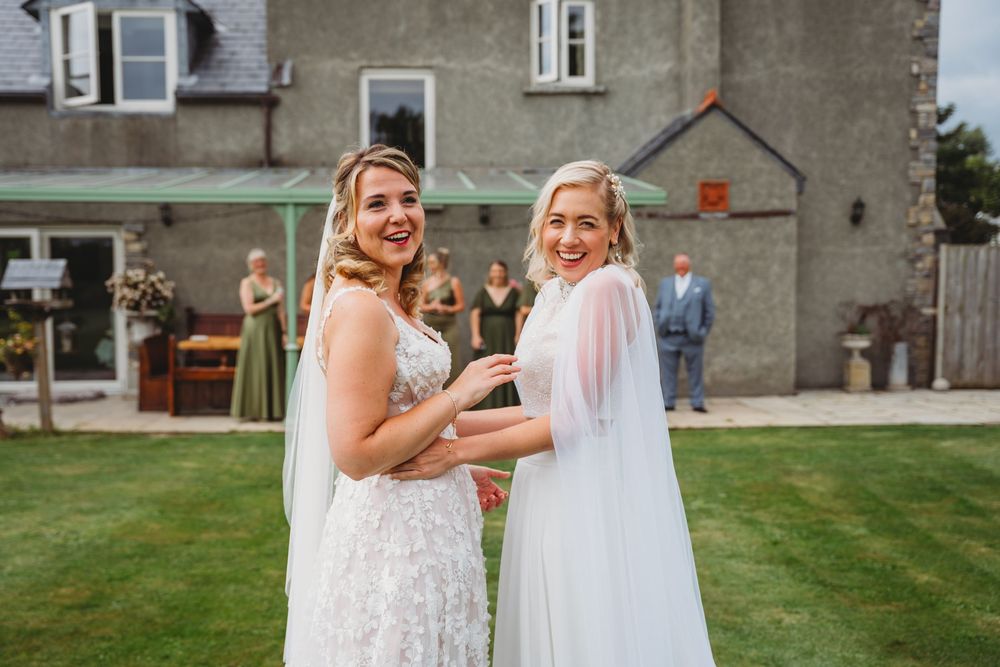 Two smiling brides in white wedding gowns embrace on a green lawn outside a stone venue, with guests in the background.