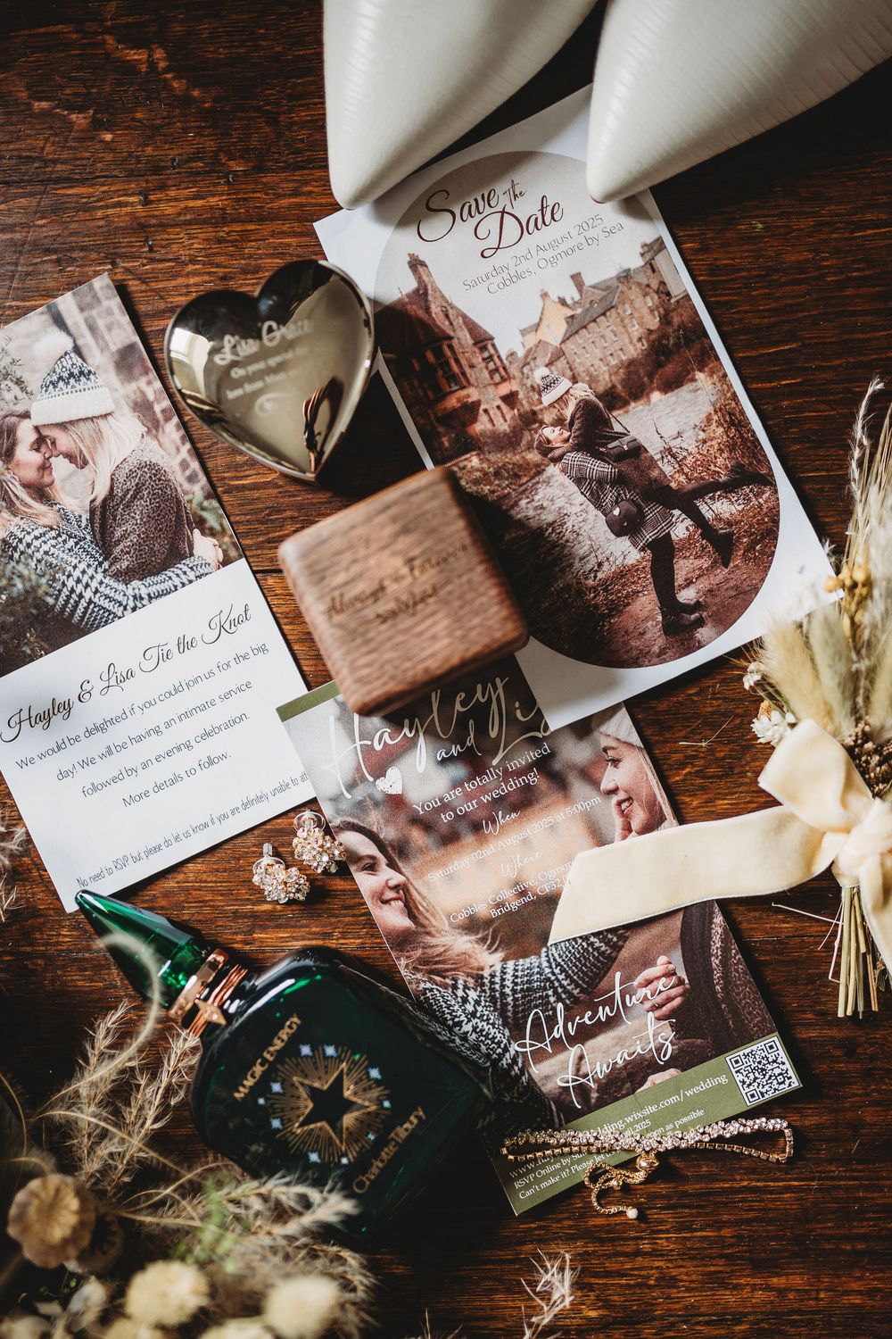 Flat lay of wedding save the date cards, a wooden ring box, heart keepsake, green bottle and dried flowers on a rustic wooden table.