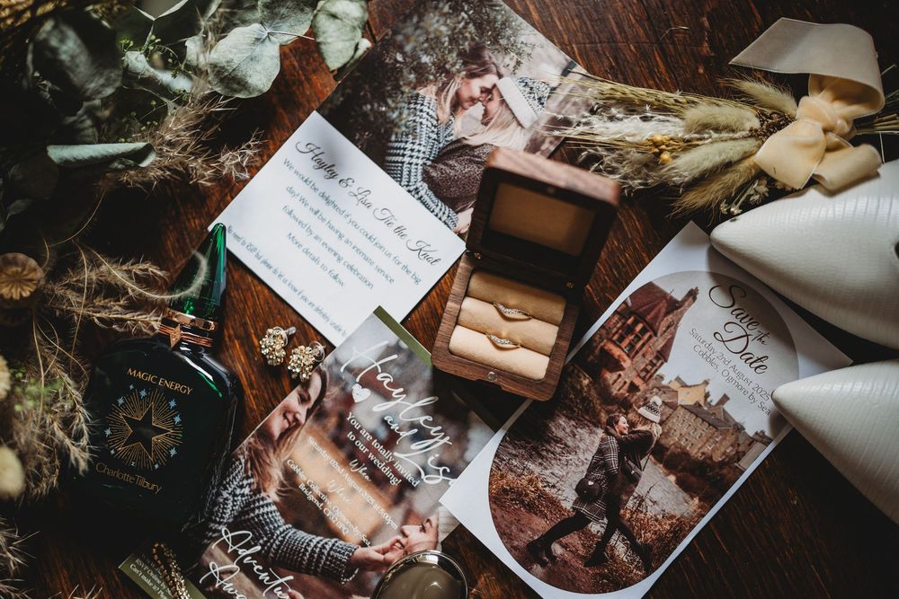 Wedding flat lay with an open ring box, printed photos, a handwritten letter, dried flowers, white heels and a perfume bottle on dark wood.
