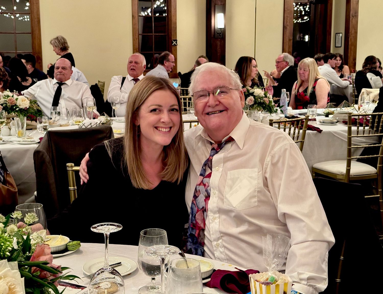 A smiling woman with long hair sits beside an older man in a patterned tie at a banquet table during a formal dinner event.