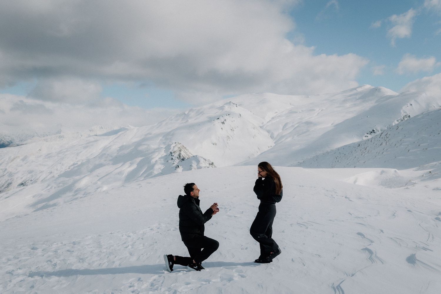 Man kneeling on one knee proposing to a woman in dark winter clothing on a snowy mountain with dramatic clouds overhead.