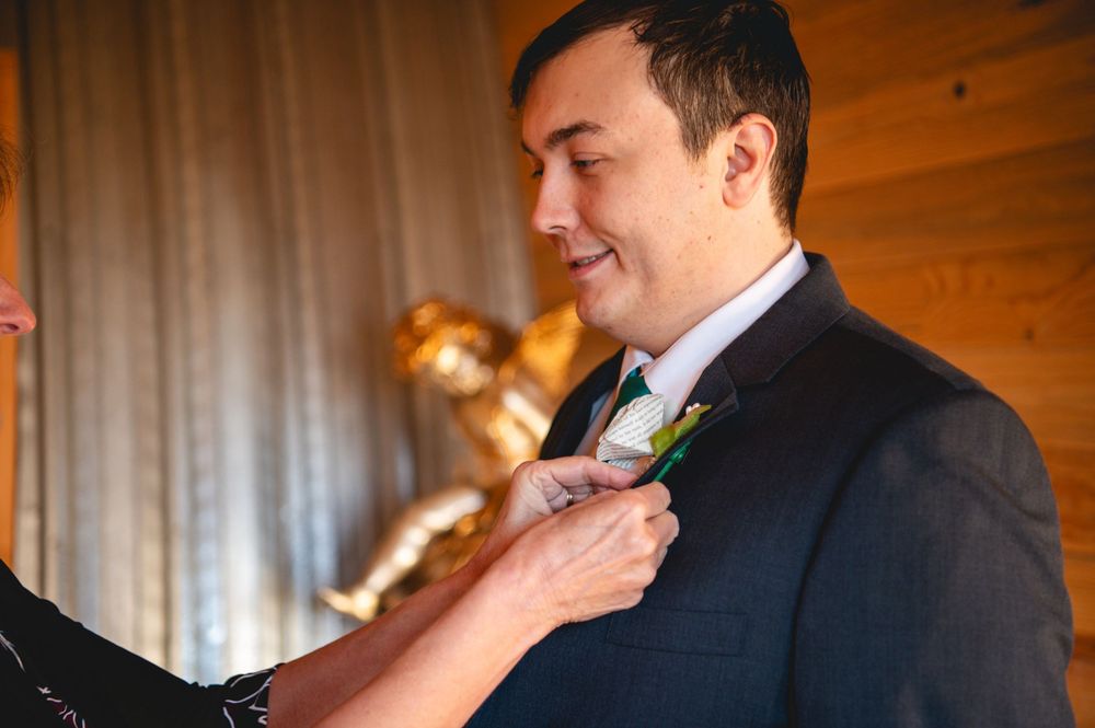 Smiling groom in a dark suit and green tie has a boutonniere pinned to his lapel in a rustic wood-paneled room.