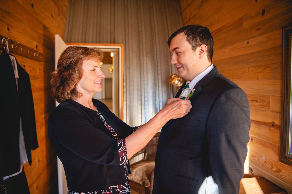 A smiling woman pins a boutonniere onto the lapel of a man in a dark suit inside a rustic wood-paneled room before a wedding.