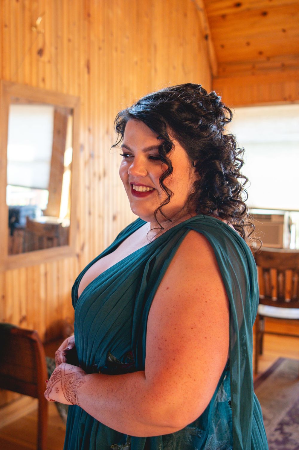 Smiling woman with dark curly updo wearing a teal green dress, standing in a rustic wood-paneled room with a mirror and wooden chairs.