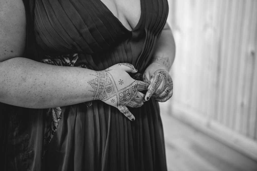 Black and white photo of a woman in a dark pleated dress clasping her hands, showing intricate henna designs on her skin.
