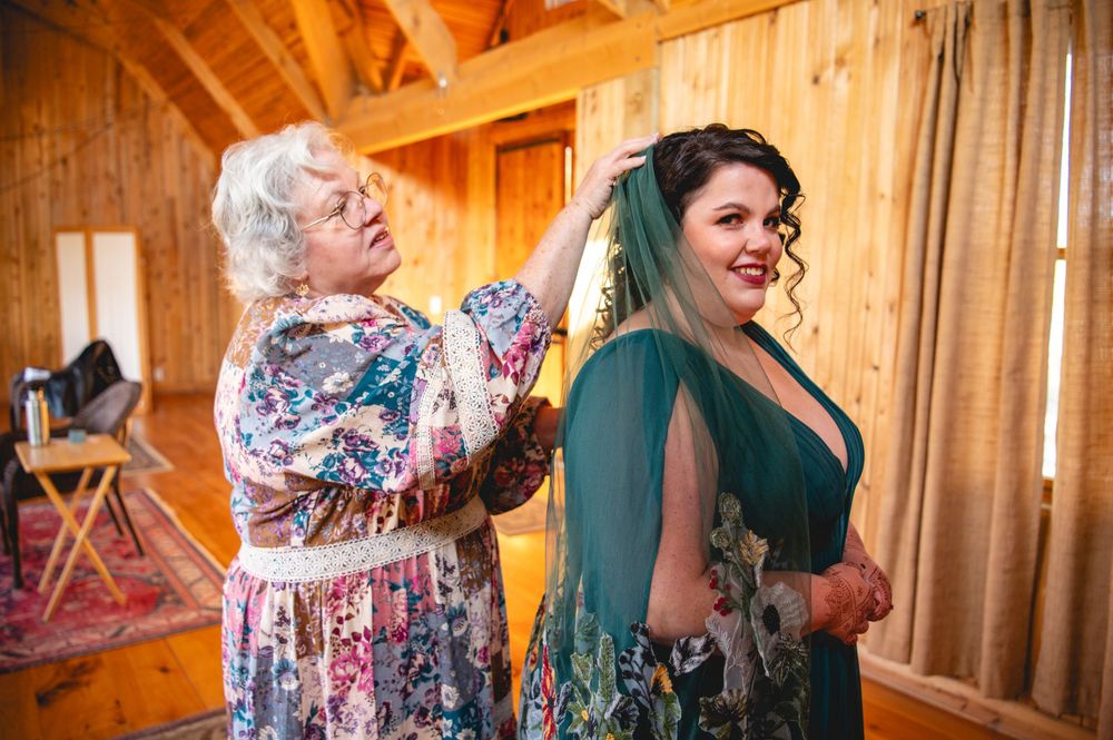 An older woman in a floral dress adjusts a smiling bride's dark curly hair and green veil inside a rustic wooden barn.
