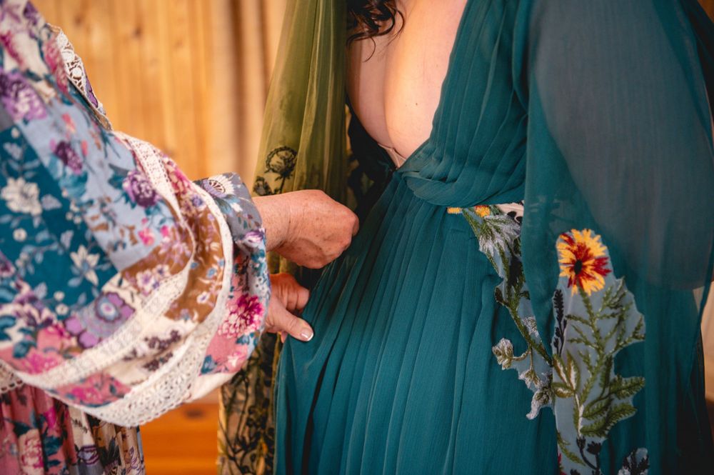 Woman in a floral kimono robe helps fasten the back of a teal green dress with embroidered flowers in a warm, softly lit room.