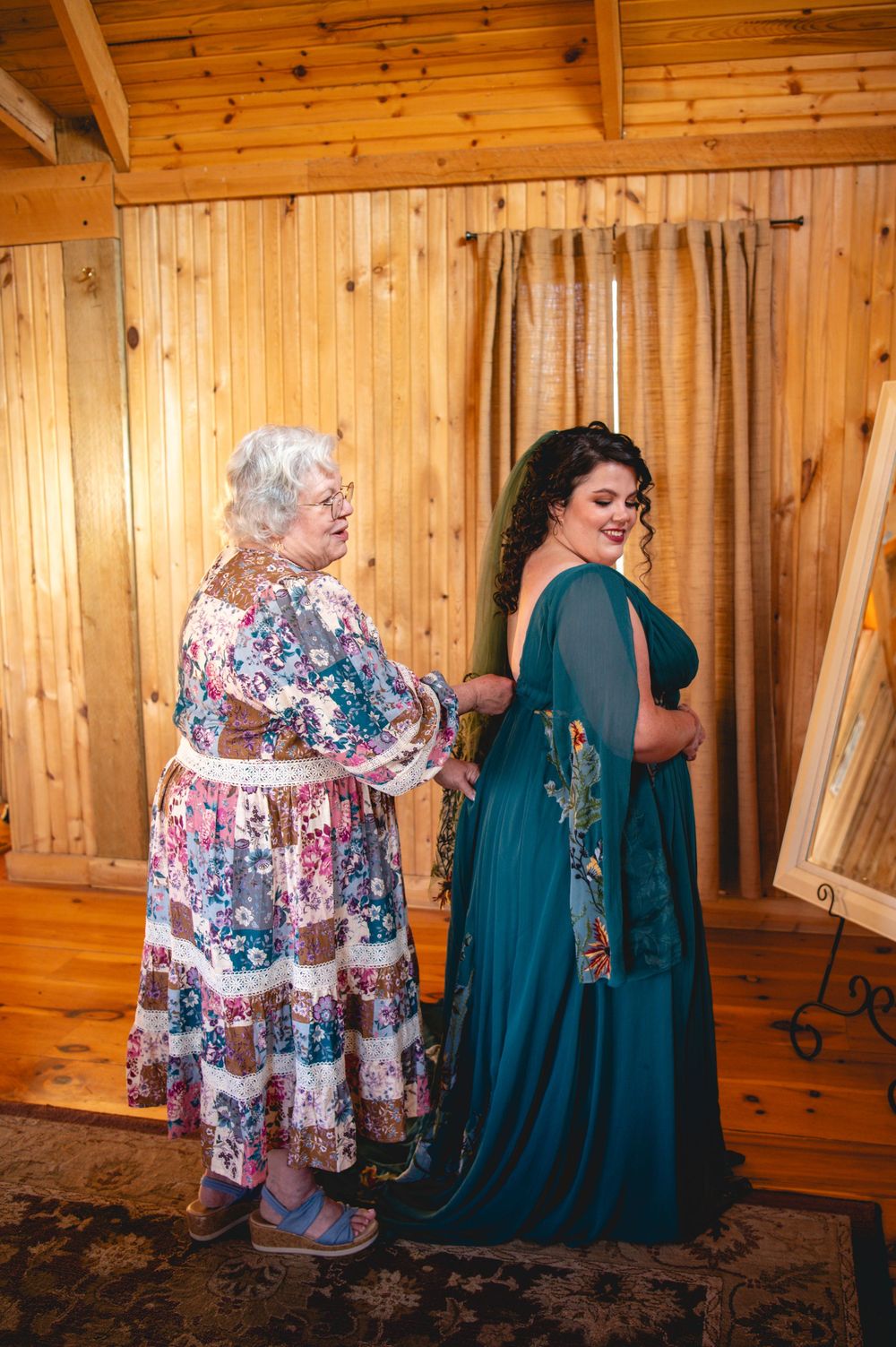 Older woman in a floral patchwork dress helps a smiling woman in an emerald green gown get ready in a rustic wood-paneled room.