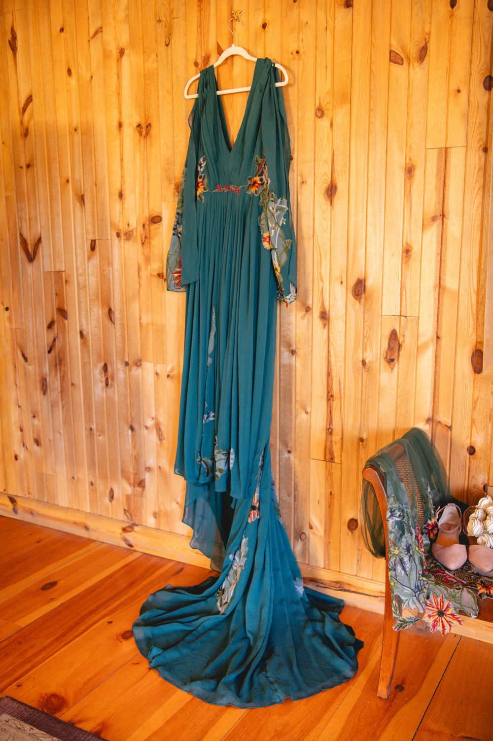 Teal embroidered boho gown with flowing train hanging on a rustic wood plank wall beside a chair holding shoes and flowers.