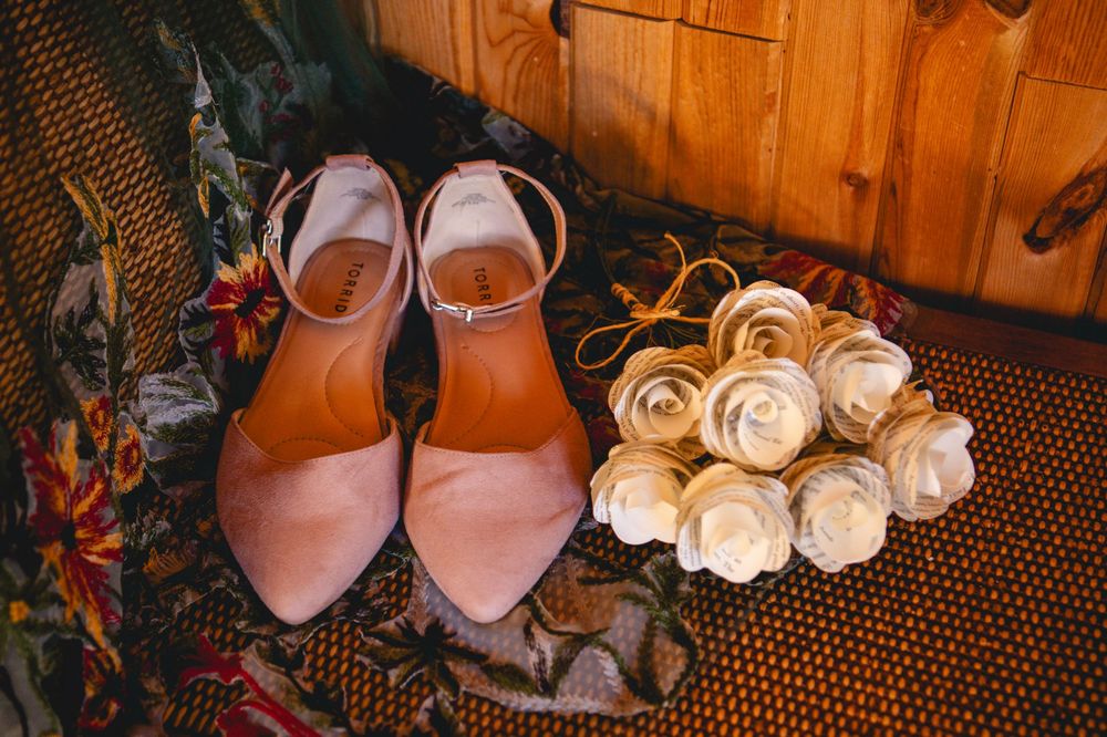 Blush pink pointed-toe wedding heels with ankle straps beside a cream paper flower bouquet on a floral rug and rustic wooden background.