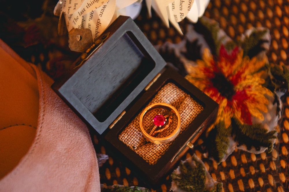 Gold ring with a red gemstone resting in an open rustic wooden box beside autumn flowers and vintage book page decor.