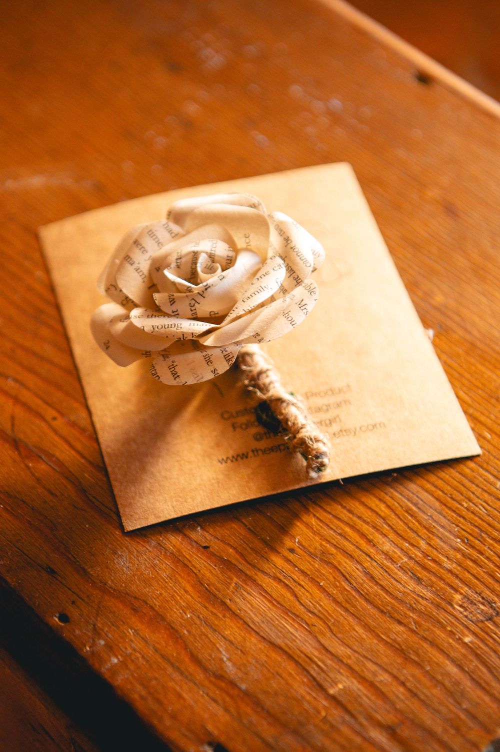 A handmade rose boutonniere made from vintage book pages with a twine-wrapped stem, resting on a kraft paper card on a wooden table.