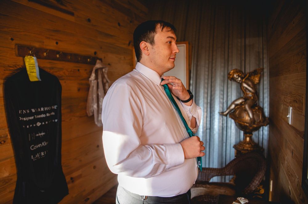 A groom in a white dress shirt adjusts his teal tie while getting ready in a rustic wood-paneled room with a statue nearby.
