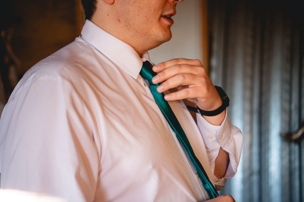 Man in a white dress shirt adjusts a teal striped necktie while getting dressed, with a watch on his wrist and curtains in the background.