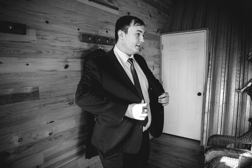 Black and white photo of a man in a dark suit and tie adjusting his cuff beside a white door in a rustic wood-paneled room.