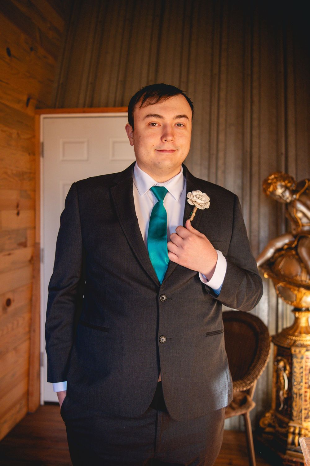 Groom in a dark suit and teal tie adjusting a white boutonniere in a rustic wood-paneled room beside an ornate gold statue.