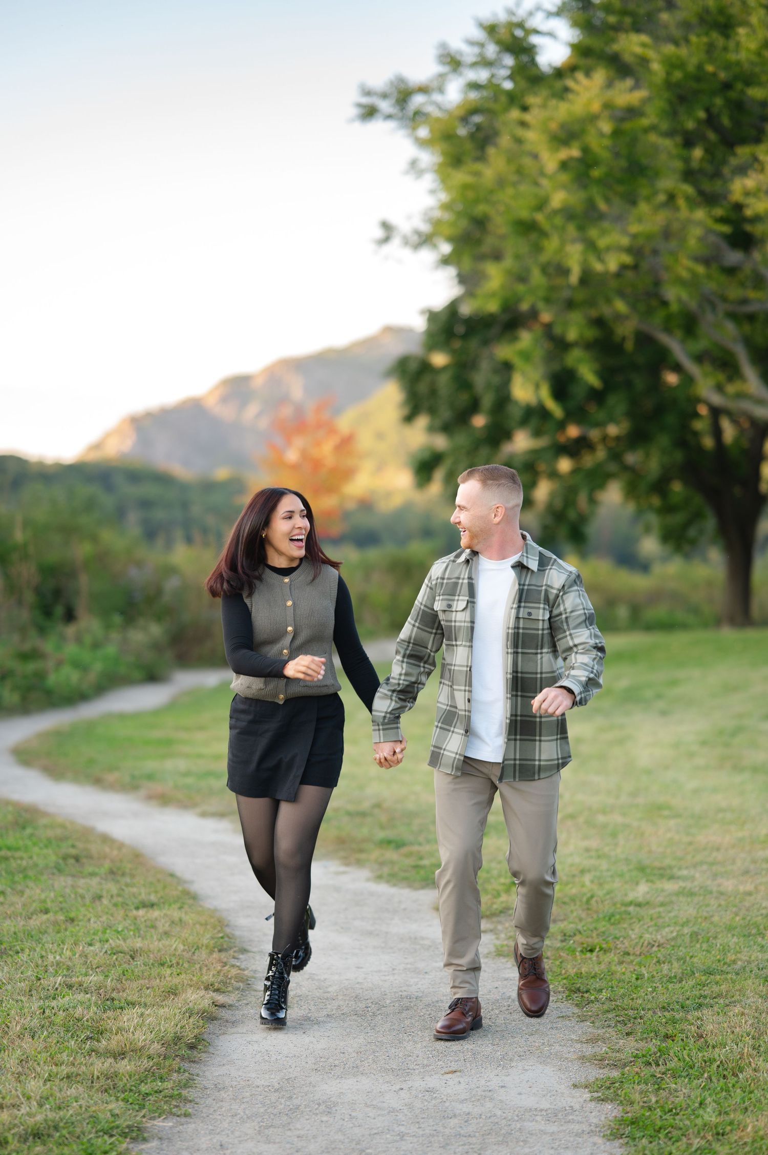 Smiling couple holding hands and running along a winding path through a grassy park with trees and mountains behind after a proposal.