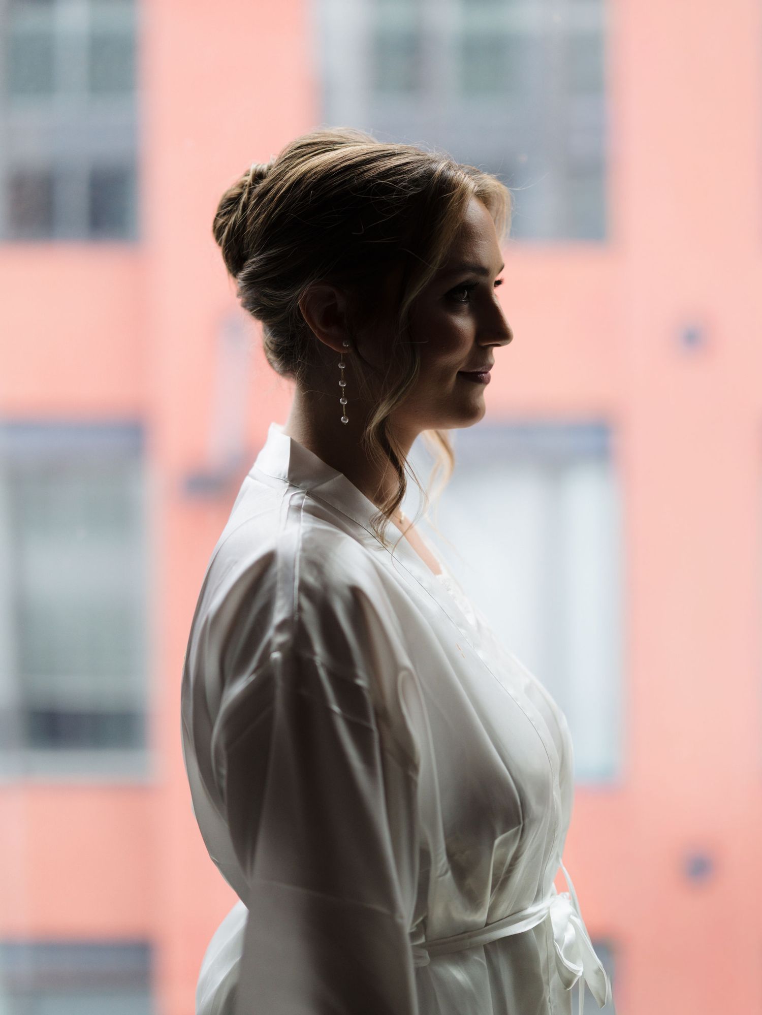 Side profile of a bride in a white satin robe with an elegant updo and drop earrings, standing by a window with a pink building behind.