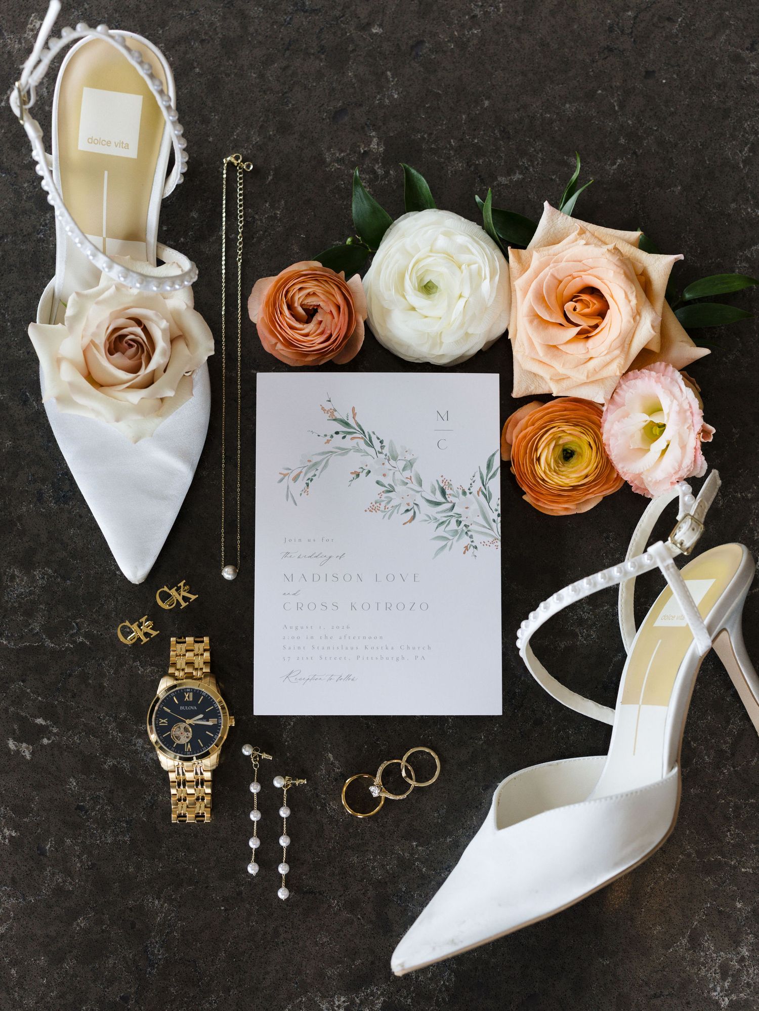 Flat lay of white satin bridal heels, a floral wedding invitation, gold watch, jewelry, and peach and cream ranunculus on a dark surface.