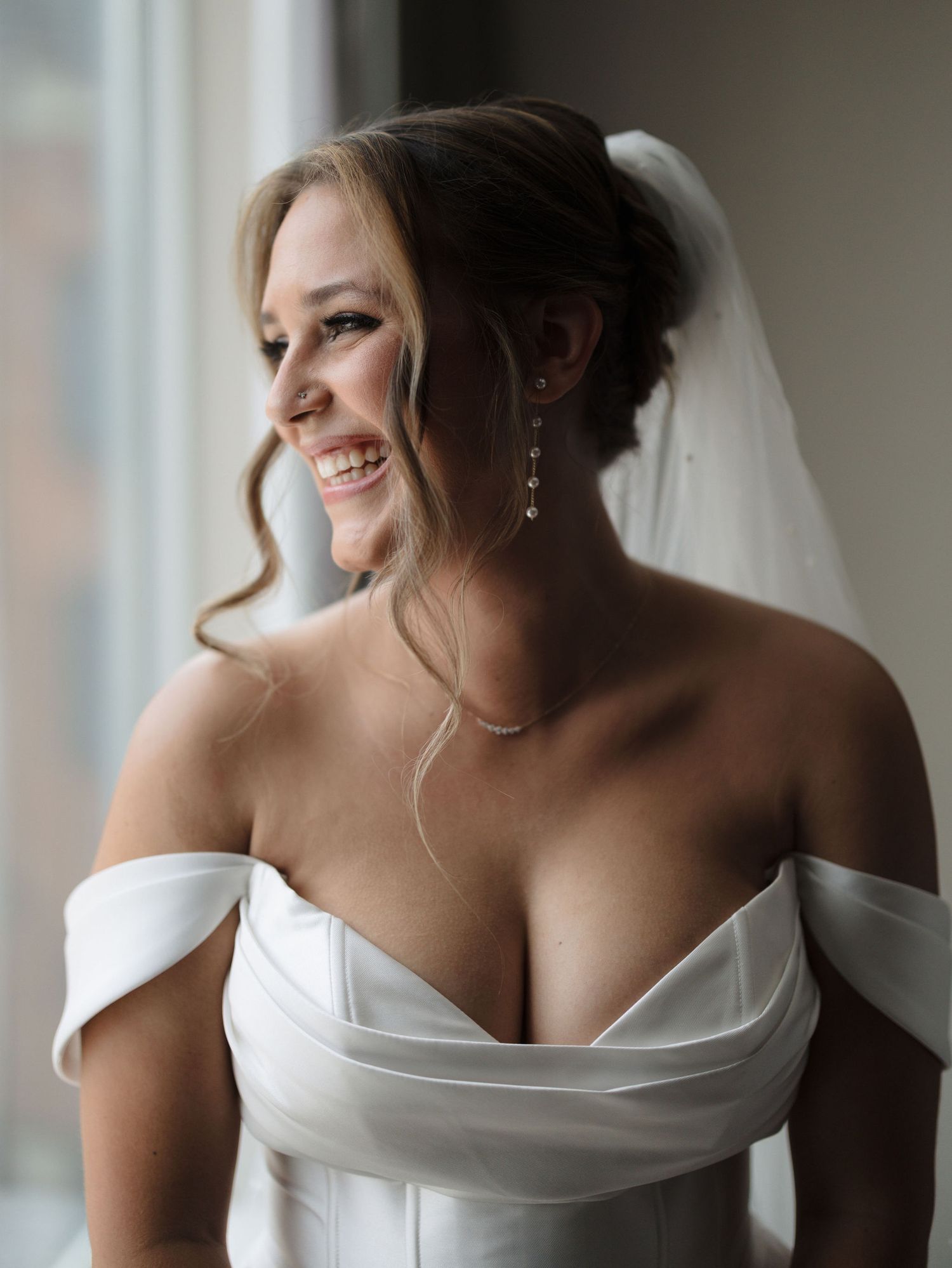 Smiling bride in an off-the-shoulder white satin wedding gown and veil gazes out a bright window with drop earrings and loose curls.