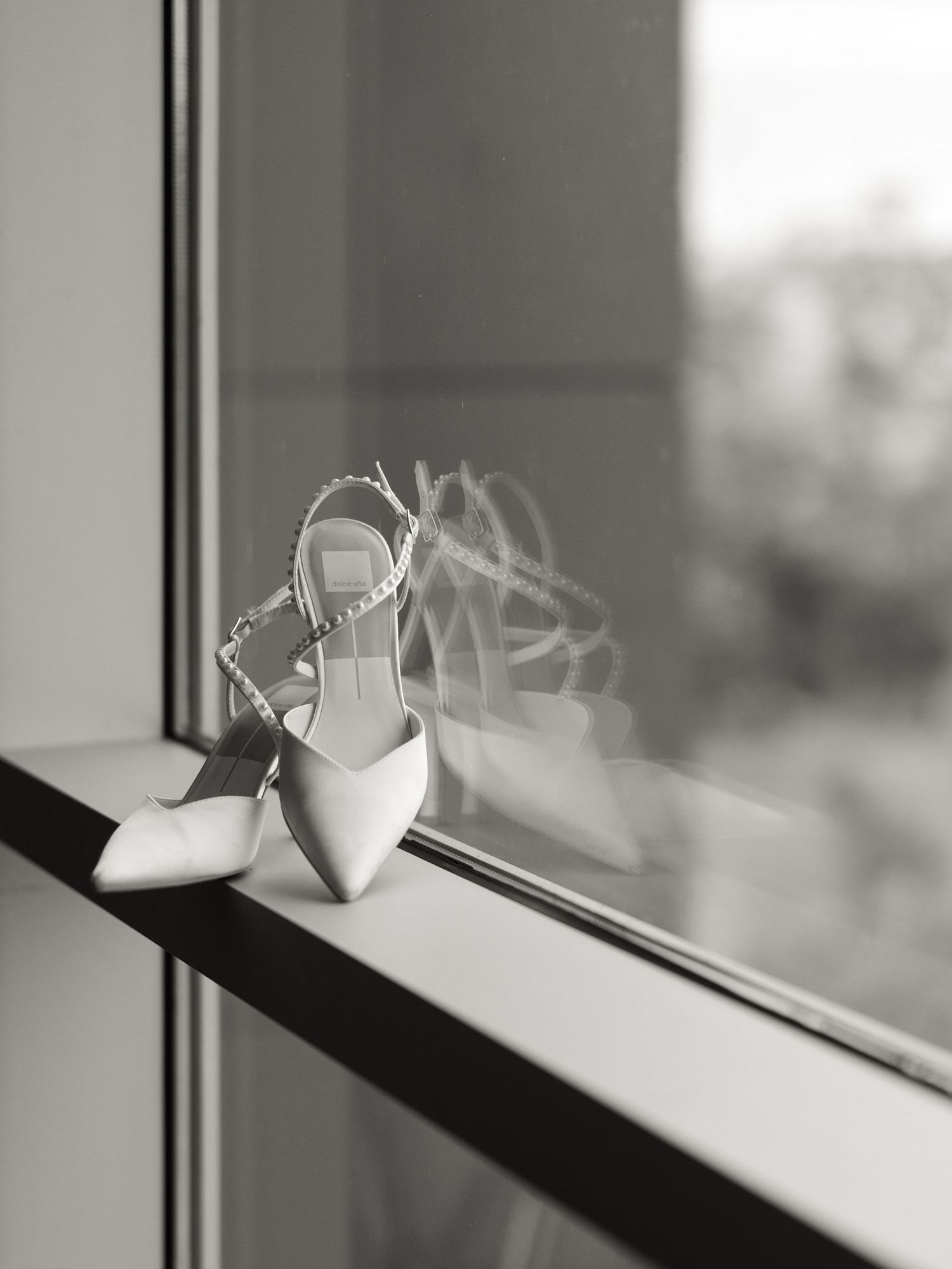 Black and white photo of pearl-strapped pointed bridal heels resting on a window ledge with a soft reflection in the glass.