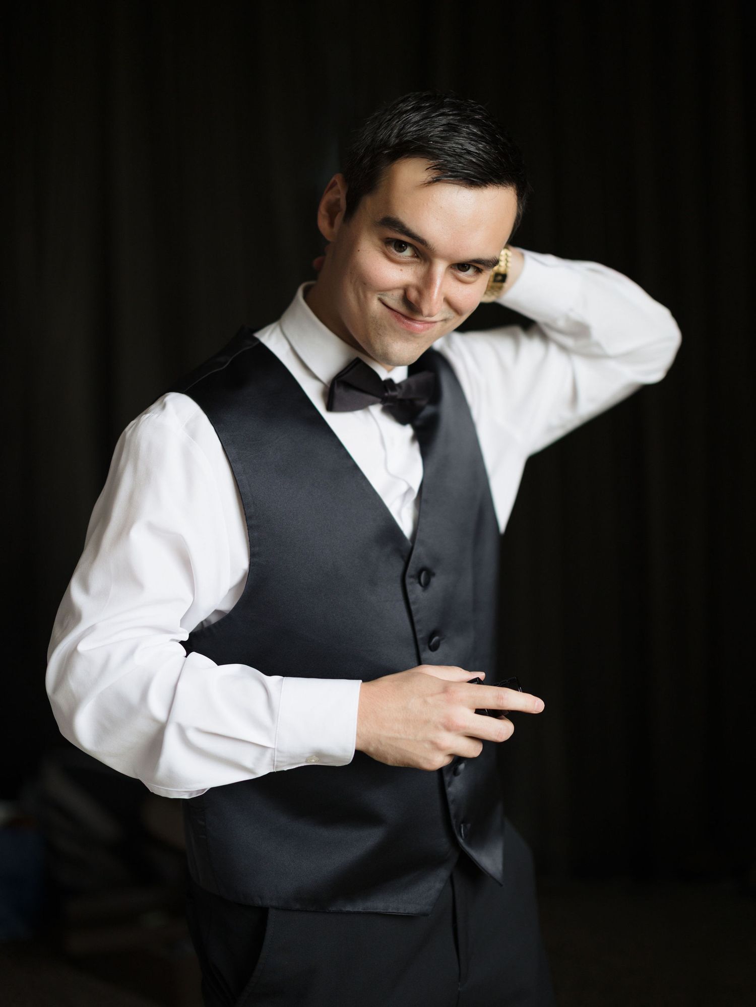 Smiling man in a black vest, white dress shirt and bow tie posing playfully with one hand behind his head against a dark backdrop.