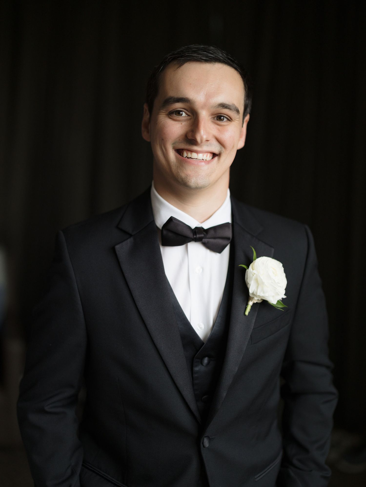 Smiling groom in a black tuxedo with a bow tie and white rose boutonniere standing against a dark curtain backdrop.