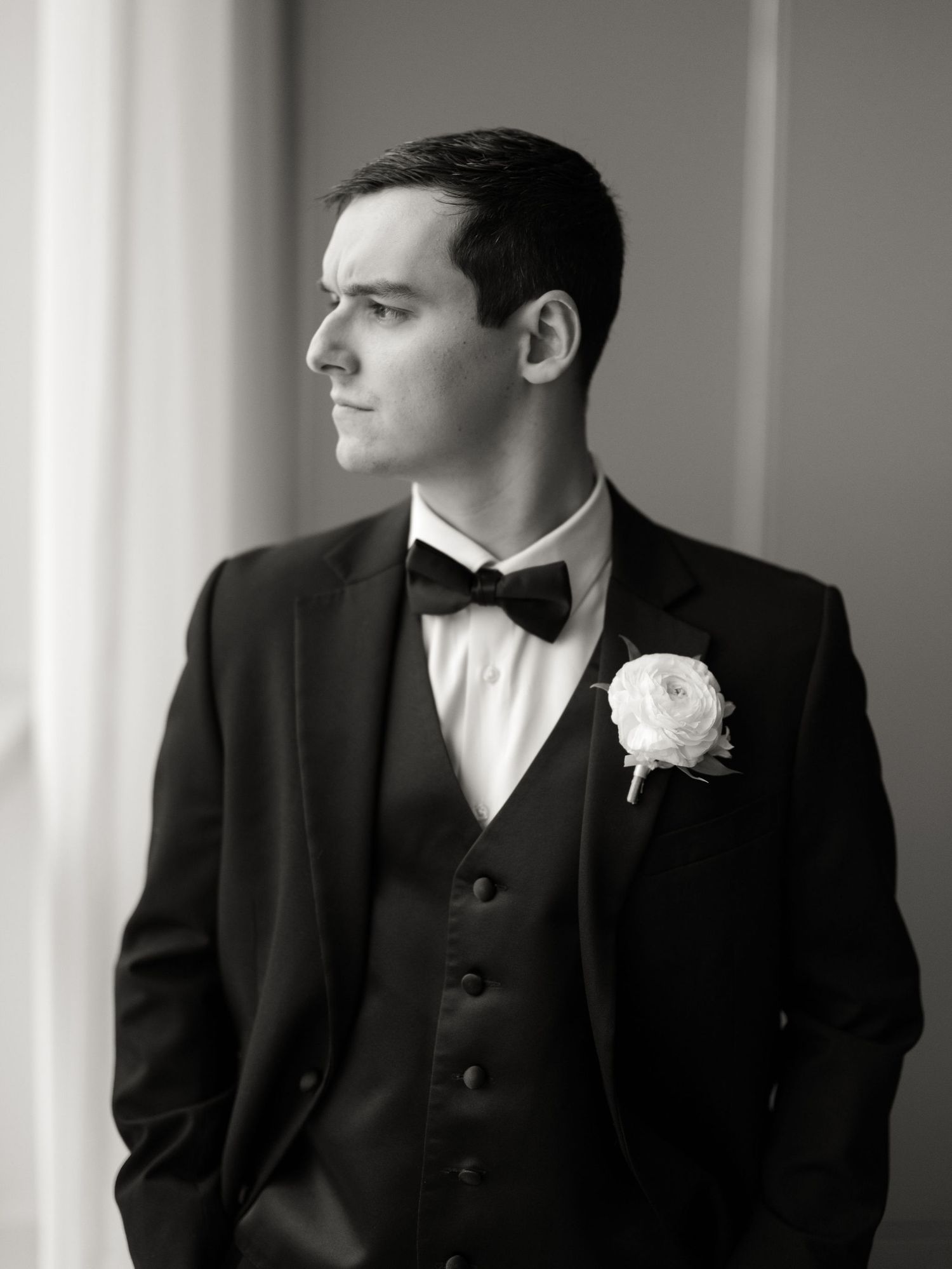 Black and white portrait of a groom in a tuxedo, bow tie and vest with a white boutonniere, gazing thoughtfully out a window.