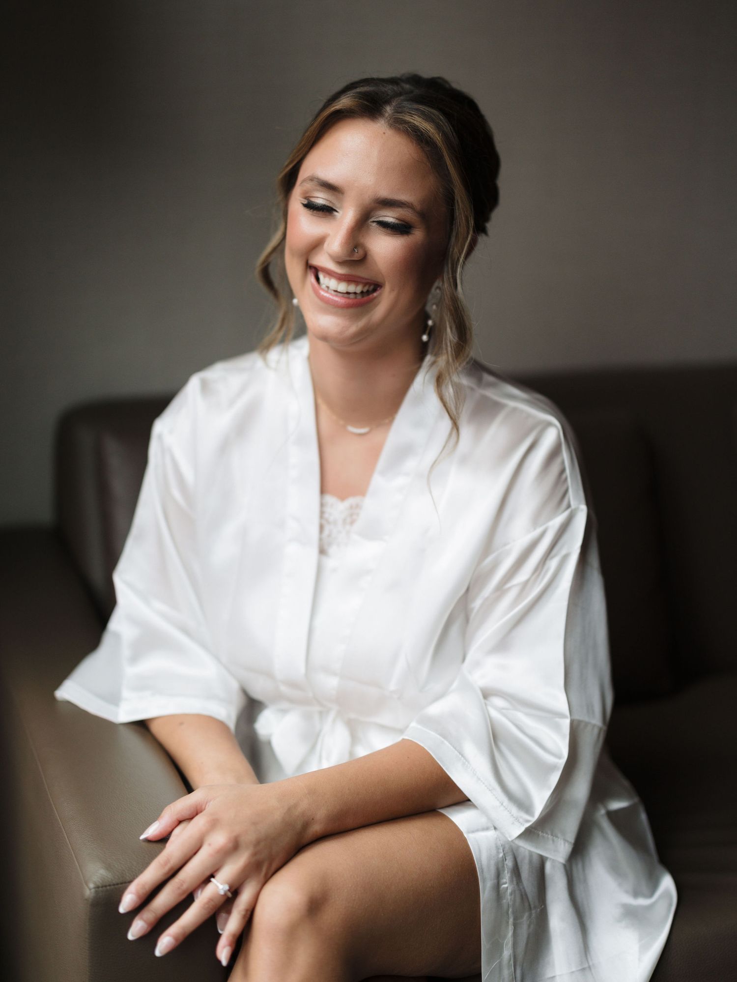 Smiling bride with an elegant updo sits on a brown leather sofa in a white satin robe, showing her ring while getting ready.
