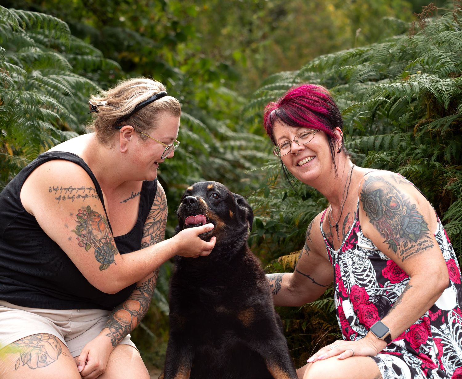 Two smiling tattooed women crouch among green ferns, petting a happy black and tan dog outdoors.