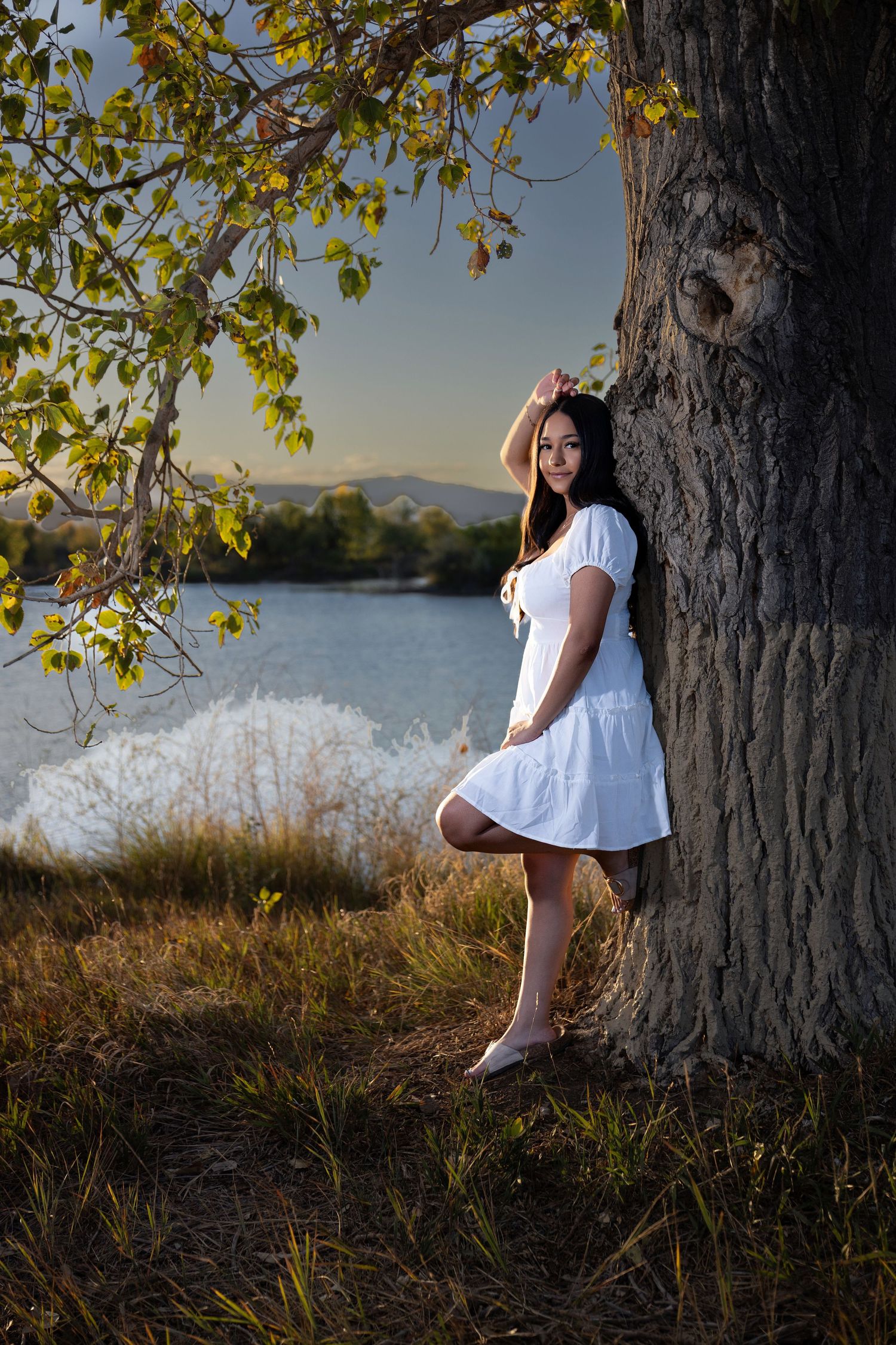 Young woman in white dress leaning against large tree by a lake at golden hour.