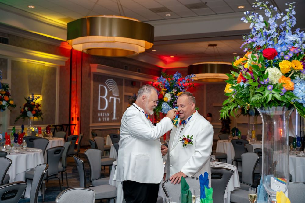 Two grooms in white suits exchange vows at an elegant wedding reception with colorful floral arrangements.