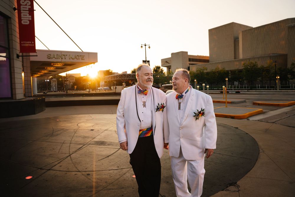 Two grooms in white suits walk together at sunset, smiling, with rainbow boutonnieres at an outdoor plaza.