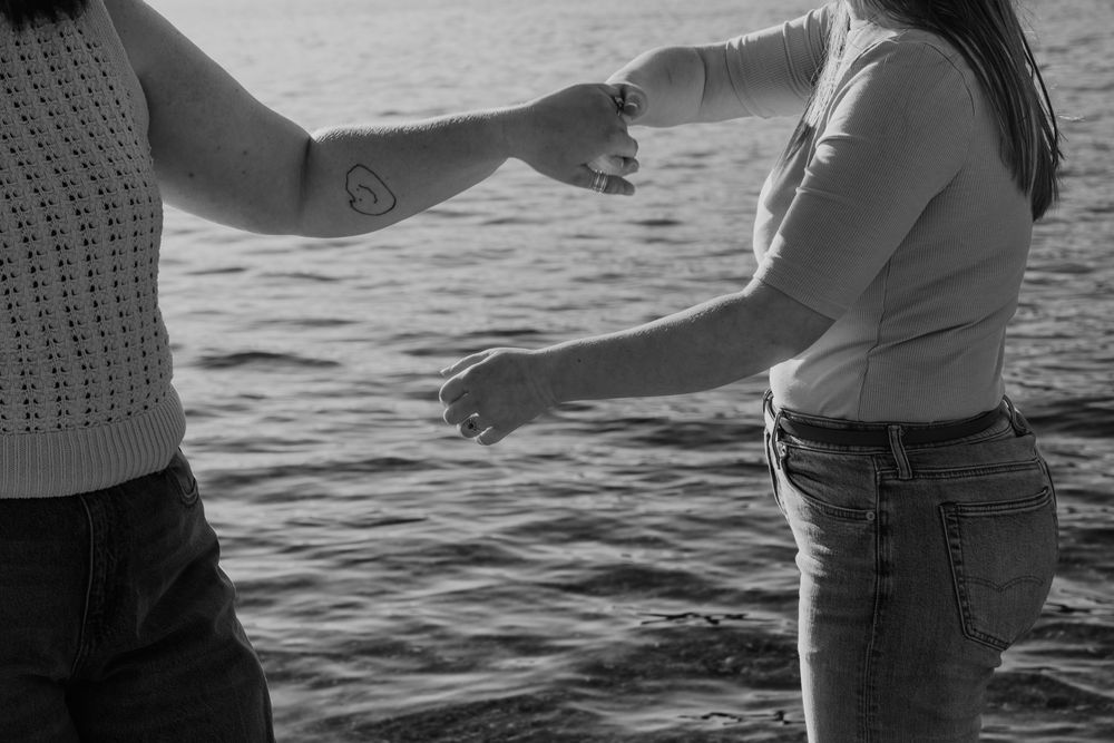 Two women holding hands near water in black and white, one with a heart tattoo on her arm.