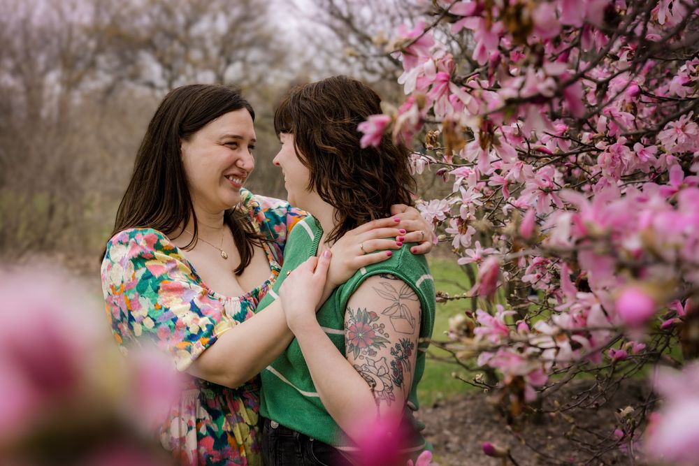 Two women share a tender moment among pink flowering trees in a romantic spring outdoor engagement photo.