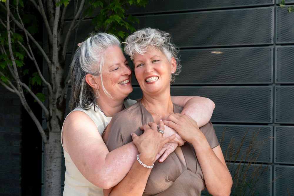 Two older women with gray hair embrace warmly outdoors, smiling joyfully near a modern building.