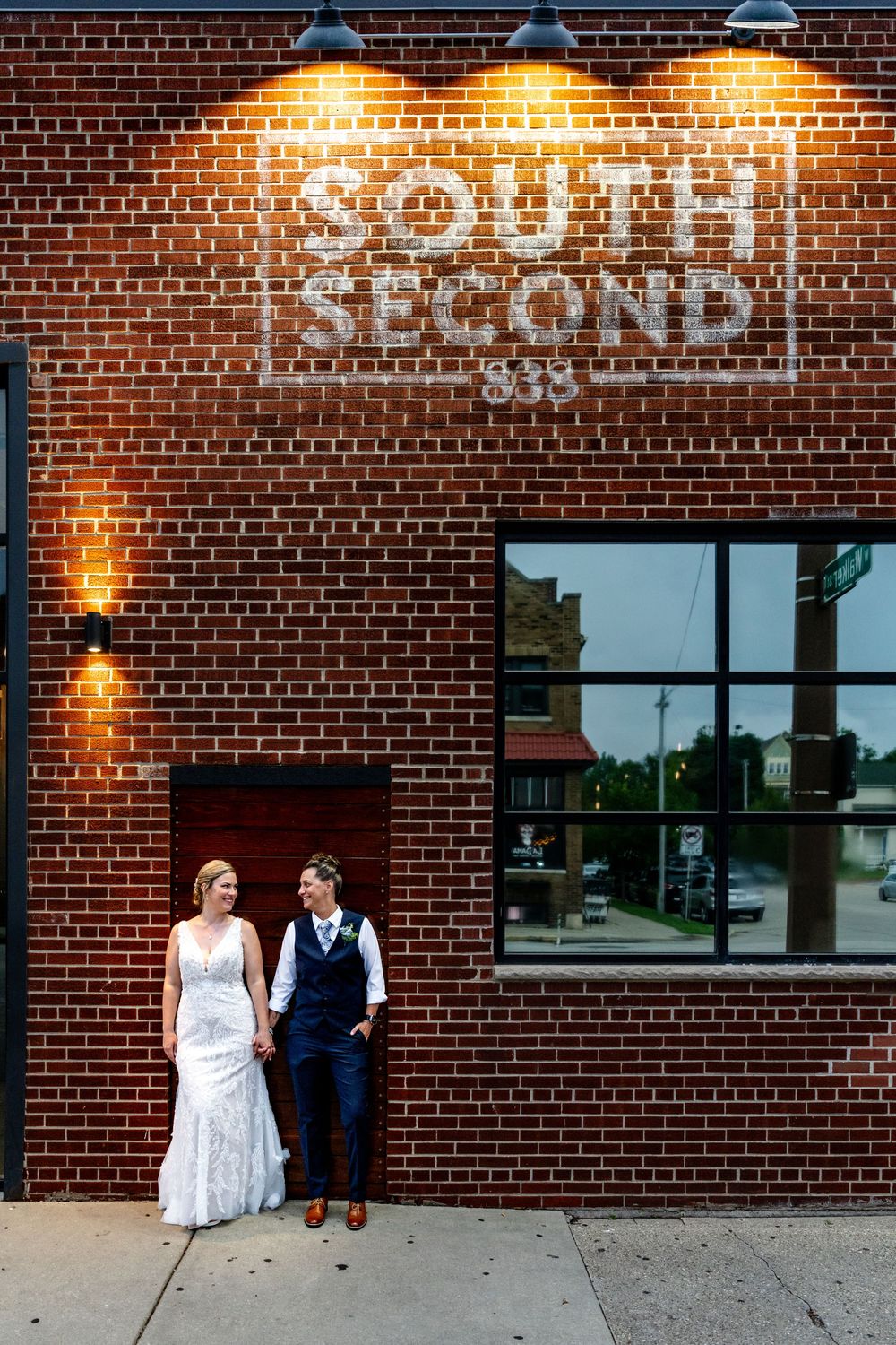 Bride and groom hold hands outside South Second brick building with glowing sign at dusk.