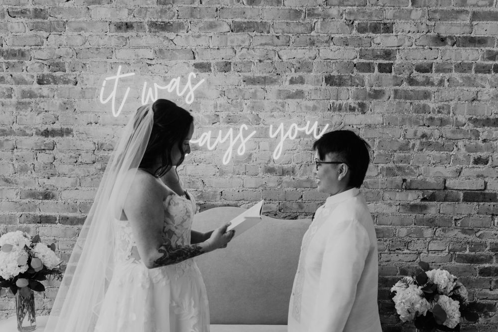 Bride and groom exchanging vows at wedding altar with neon sign and brick wall backdrop.