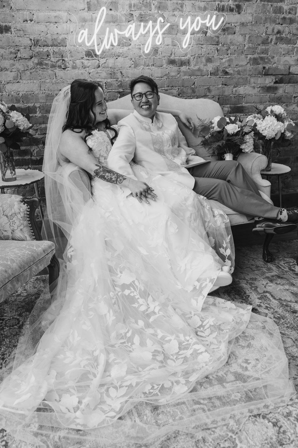 Black and white wedding photo of a laughing couple sitting on a vintage sofa with an always you neon sign above.