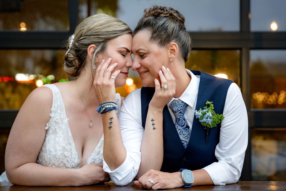 Two brides share an intimate moment, touching foreheads and smiling, wearing wedding attire with floral boutonniere.