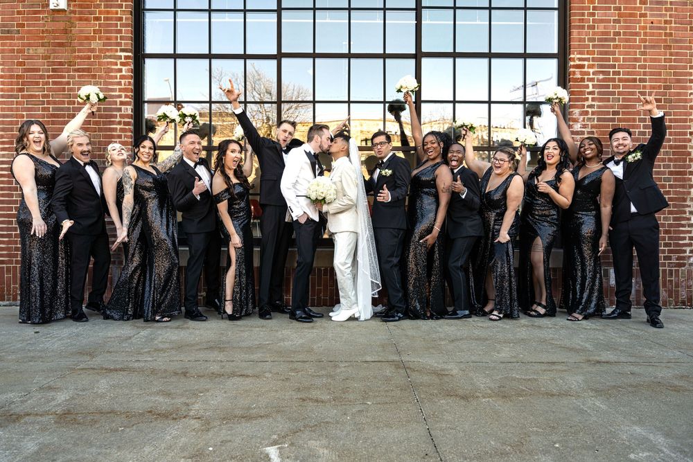 Two grooms kiss surrounded by wedding party in black attire, celebrating in front of brick wall.